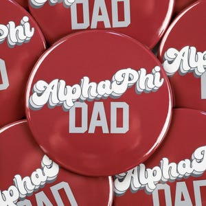 May include: A close-up of several red round buttons with white text that reads "Alpha Phi Dad" in a retro font.