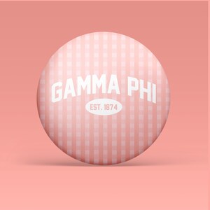 May include: A circular pinback button featuring a light pink and white gingham pattern. The text GAMMA PHI is printed in white in an arched font above a white oval containing the text EST. 1874 in light pink, centered on the button surface.