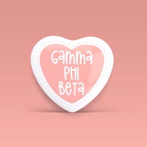 May include: A heart-shaped pin with a glossy light pink center and a white border featuring a dotted pattern. The text Gamma Phi Beta is printed in white, stylized, rounded lettering across the center of the heart-shaped surface.