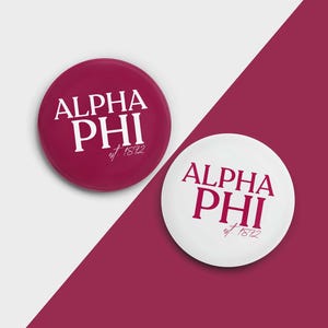 May include: Two round buttons with the text "ALPHA PHI est. 1872" in white lettering. One button is red and the other is white.