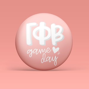 May include: A circular pinback button with a matte light pink surface. The center features the white Greek letters Gamma Phi Beta, followed by the white cursive text game day and a small white heart icon positioned to the right of the word game.