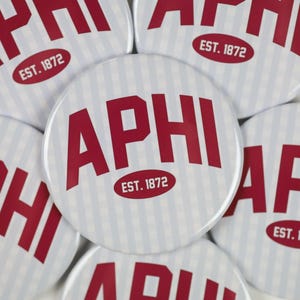 May include: A collection of round buttons with the letters "APHI" in red, centered on a white background with vertical light blue stripes. Each button also features the text "EST. 1872" in a red oval.