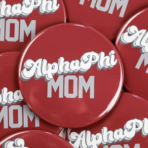 May include: A close-up of several red round buttons with the text "Alpha Phi MOM" in white and gray lettering. The buttons are arranged in a pile.