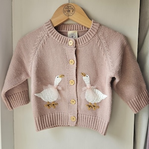 May include: A dusty pink knit cardigan featuring a crew neckline, long sleeves, and five light-coloured buttons. The front displays two embroidered white geese with tulle accents and yellow feet. A branded label is visible at the inner neckline.