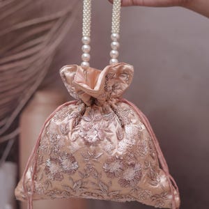 Puede incluir: Un bolso con cordón bordado de color melocotón con un asa de perlas. El bolso presenta bordados florales y detalles de lentejuelas. Los cordones terminan en borlas de perlas. El bolso está sostenido por una mano.
