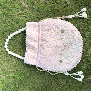 Puede incluir: Un bolso de cord&oacute;n rosa con cuentas, con un asa de perlas y un borla. El bolso est&aacute; decorado con bordados blancos y dorados.
