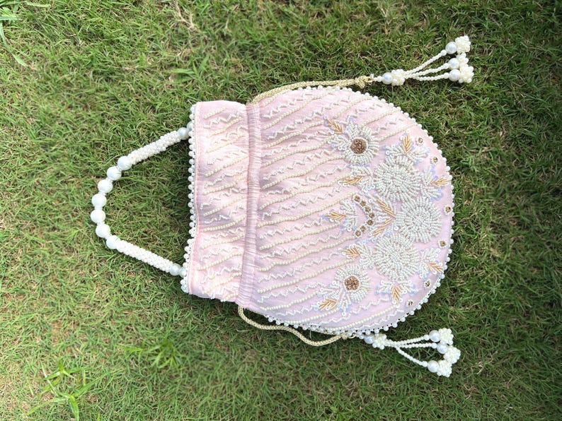 Puede incluir: Un bolso de cord&oacute;n rosa con cuentas, con un asa de perlas y un borla. El bolso est&aacute; decorado con bordados blancos y dorados.