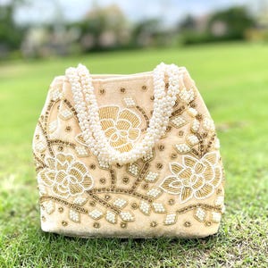 Puede incluir: Un bolso de mano de terciopelo crema con un diseño floral bordado con cuentas doradas y blancas. El bolso tiene una correa de perlas.