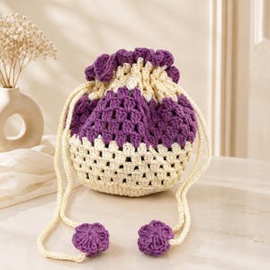 May include: A small, round, crocheted drawstring pouch featuring a cream-colored base and a purple upper section. The bag has a ruffled purple top edge, cream-colored drawstrings, and two small, purple, flower-shaped accents at the ends.