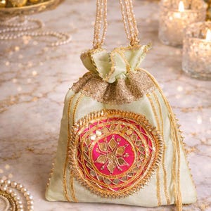 May include: A light green drawstring pouch with gold and pink embroidery. The pouch features a central pink medallion with gold mirror accents and a gold floral design. The handle is made of beaded strands. The pouch is decorated with gold trim.