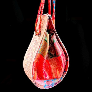 Könnte beinhalten: Eine farbenfrohe, handgefertigte Tragetasche mit einzigartigem Design. Die Tasche hat eine weiße, spitzenartige Außenseite und ein gemustertes Innenfutter mit roten, grünen und blauen Akzenten. Die Tasche hat rote Träger und Metallösen.