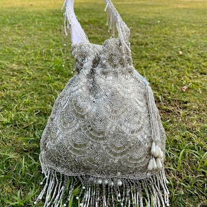 Puede incluir: Un bolso de cord&oacute;n de cuentas plateadas con flecos y detalles de perlas. El bolso tiene un asa de tela blanca y est&aacute; fotografiado sobre una superficie de hierba verde.