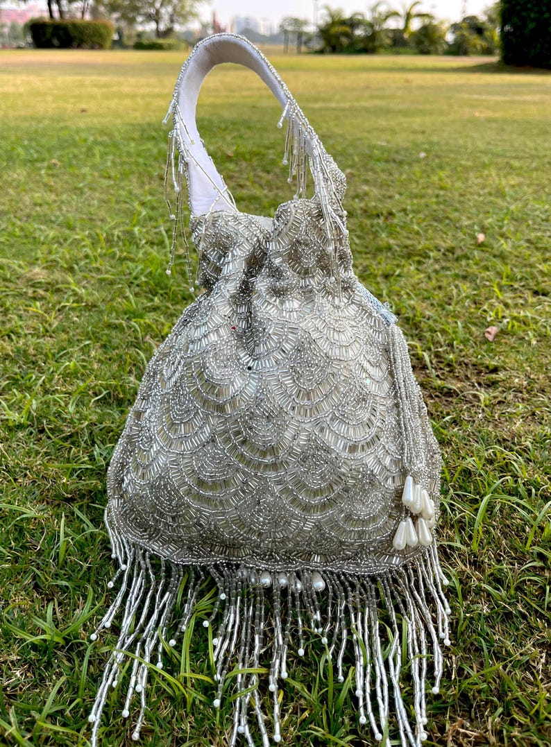 Puede incluir: Un bolso de cord&oacute;n de cuentas plateadas con flecos y detalles de perlas. El bolso tiene un asa de tela blanca y est&aacute; fotografiado sobre una superficie de hierba verde.