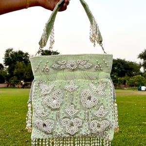 May include: A light green velvet handbag with a beaded floral design and fringe. The bag has a long strap and a drawstring closure.