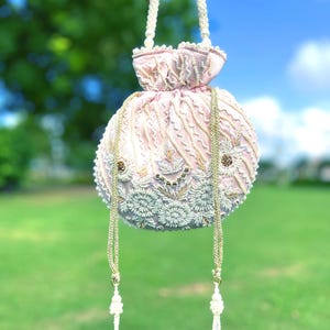 Puede incluir: Un bolso de cord&oacute;n rosa con cuentas, con un asa de perlas blancas y borlas. El bolso est&aacute; decorado con bordados dorados y blancos.