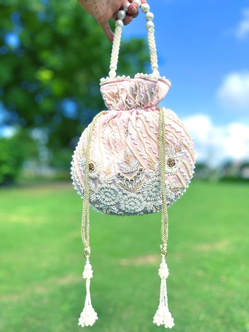 Puede incluir: Un bolso de cord&oacute;n rosa con cuentas, con un asa de perlas blancas y borlas. El bolso est&aacute; decorado con bordados dorados y blancos.