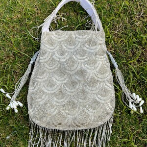 Puede incluir: Un bolso de mano de cuentas plateadas con ribete de flecos. El bolso tiene un asa de tela blanca y un dise&ntilde;o de escamas.