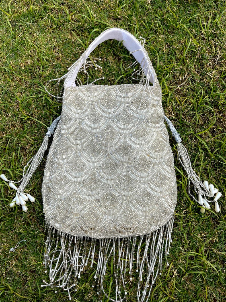 Puede incluir: Un bolso de mano de cuentas plateadas con ribete de flecos. El bolso tiene un asa de tela blanca y un dise&ntilde;o de escamas.