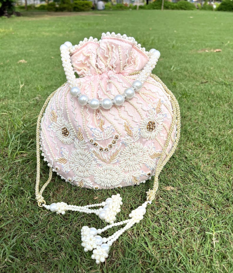 Puede incluir: Un bolso de cord&oacute;n rosa con cuentas, con una correa de cadena dorada y un borla. El bolso est&aacute; decorado con bordados blancos y dorados.