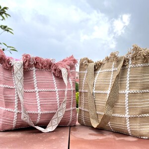 Pode incluir: Duas sacolas de tecido trançado com detalhes em franjas. Uma sacola é rosa com listras brancas e a outra é bege com listras brancas.