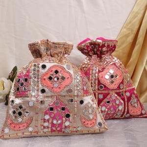 May include: Two decorative drawstring bags. One is beige with pink and silver embellishments, the other is red with pink and silver accents. Both bags feature mirrored details and intricate embroidery, creating a festive and ornate appearance.