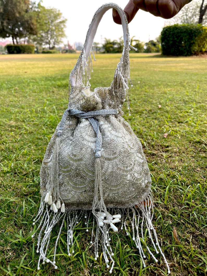 Puede incluir: Un bolso de cord&oacute;n de cuentas plateadas con flecos largos. El bolso tiene un forro de sat&eacute;n blanco y un cierre de cord&oacute;n plateado.