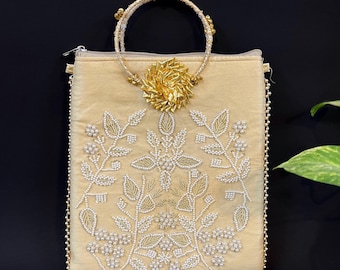 May include: A beige clutch bag with a gold ring handle and a floral design embroidered with white beads. The bag has a gold zipper closure and a gold chain strap.