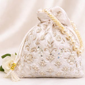 Könnte beinhalten: Elfenbeinfarbener Beutel mit aufwendiger silberner Stickerei, Pailletten und einem perlenbesetzten Griff. Die Tasche hat ein Blumenmuster und eine Quaste mit goldenen Akzenten. Eine weiße Rose liegt neben der Tasche und verleiht Eleganz.