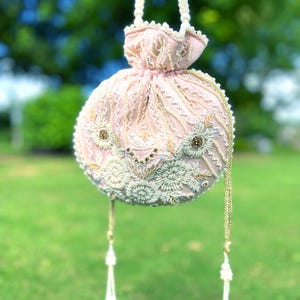 Puede incluir: Un bolso de mano rosa con cuentas, con un dise&ntilde;o floral blanco y dorado y un borla blanca. El bolso est&aacute; colgado de una correa de cuentas blancas.