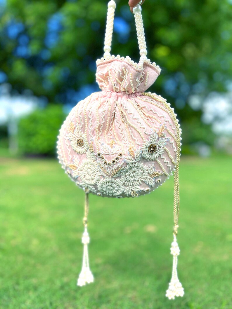 Puede incluir: Un bolso de mano rosa con cuentas, con un dise&ntilde;o floral blanco y dorado y un borla blanca. El bolso est&aacute; colgado de una correa de cuentas blancas.