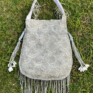 Puede incluir: Un bolso blanco de cuentas con un borde de flecos y un asa de tela blanca. El bolso est&aacute; decorado con un patr&oacute;n de peque&ntilde;as cuentas blancas.