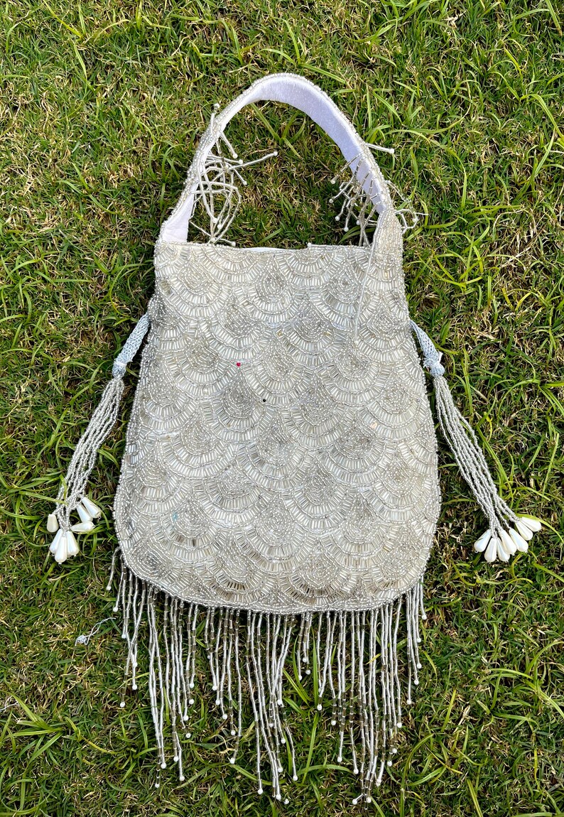 Puede incluir: Un bolso blanco de cuentas con un borde de flecos y un asa de tela blanca. El bolso est&aacute; decorado con un patr&oacute;n de peque&ntilde;as cuentas blancas.