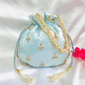 May include: A pale blue satin drawstring bag with gold embroidery and pearl detailing. The bag has a beaded handle and tassel features. The design includes gold floral patterns and small pearl embellishments, creating an elegant accessory.