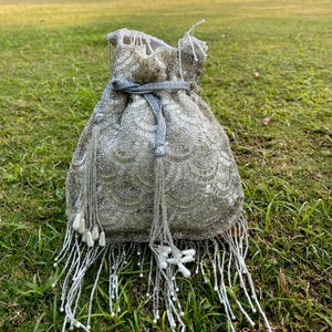 Puede incluir: Una bolsa de cord&oacute;n de cuentas plateadas con flecos de cuentas blancas. La bolsa tiene un dise&ntilde;o de conchas y un cord&oacute;n de color gris.