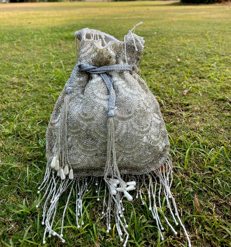 Puede incluir: Una bolsa de cord&oacute;n de cuentas plateadas con flecos de cuentas blancas. La bolsa tiene un dise&ntilde;o de conchas y un cord&oacute;n de color gris.