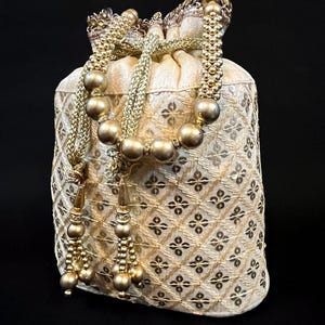 Puede incluir: Una bolsa de cordón de color dorado con un patrón geométrico y ribete de cuentas. La bolsa tiene un borla a cada lado.