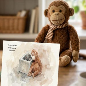 May include: A soft brown monkey toy with a striped scarf is positioned behind a watercolour print. The print shows two monkeys, one in a box, with the text "Lean on me When you Need to".