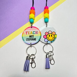 May include: A rainbow lanyard with a round tag that says "Teach Miss Stephanie" and a smiley face flower charm. The lanyard has a purple tassel and a silver clip.