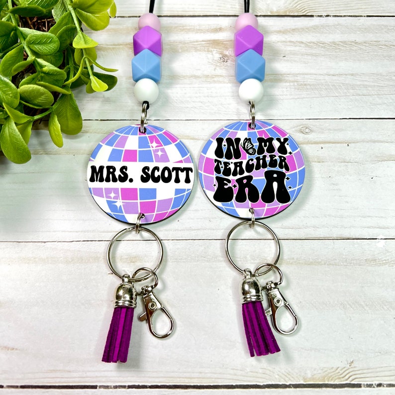 May include: Two teacher lanyards with a disco ball design in purple, blue, and white. One says "MRS. SCOTT" and the other says "IN MY TEACHER ERA". Each has a purple tassel and a silver clasp. The lanyards have a beaded necklace.