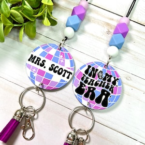 May include: Two round lanyards with a disco ball design in shades of purple, blue, and white. One says "MRS. SCOTT" and the other says "IN MY TEACHER ERA". Each has a purple tassel and a beaded lanyard.