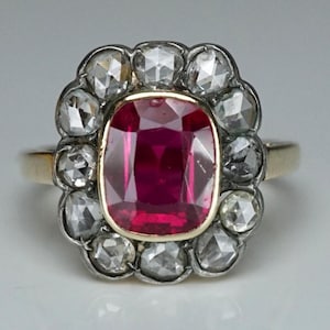 Könnte beinhalten: Ein Goldring mit einem zentralen, kissenförmigen, tiefroten Rubin. Der Rubin ist von einem Kranz aus zwölf facettierten, klaren Diamanten umgeben, die in einer dunklen Metallfassung sitzen. Der Ring besteht aus poliertem Gold und ist mittig gefasst.