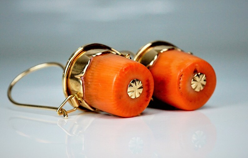 Puede incluir: Un par de pendientes de coral naranja con detalles dorados. Los pendientes tienen forma de cilindro y cierre de gancho dorado.