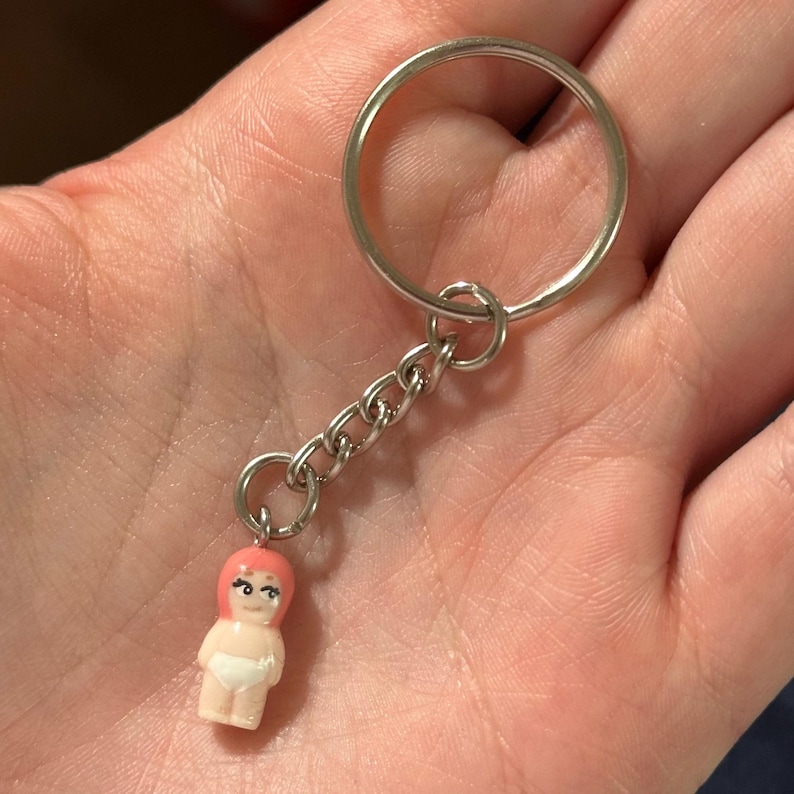 May include: A keychain with a miniature doll charm. The doll has pink hair, a white diaper, and a smiling face. The charm is attached to a silver chain and keyring.