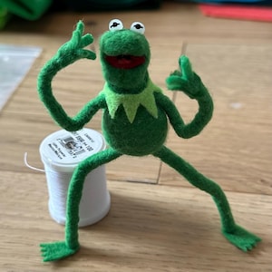 以下が含まれることがあります： 遊び心のあるポーズの、手作りのフェルト製カーミット・ザ・フロッグの置物。腕を上げ、足を広げています。緑色のカエルは白い目、赤い口、薄緑色の襟をしています。近くには白い糸巻きがあります。