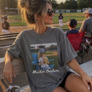 Może przedstawiać: Szara koszulka z grafiką dziewczyny na stadionie baseballowym, tekst brzmi "Bleacher Creature". Osoba nosi okulary przeciwsłoneczne i siedzi na ławce na boisku do baseballu.