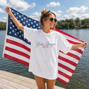 May include: A white t-shirt with the text "Your Design HERE" in a script font, held by a person in front of an American flag. The flag has red and white stripes and blue field with white stars. The background is a lake and sky.