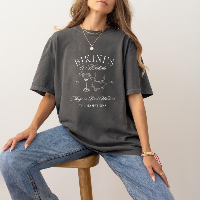 May include: A dark gray t-shirt with a white graphic design that says "Bikini's & Martinis Est. 2021 Morgan's Bach Weekend The Hamptons". The graphic includes a martini glass and a bikini.