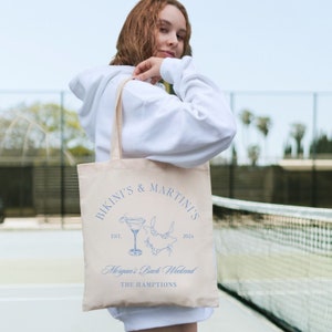 以下が含まれることがあります： ベージュのトートバッグに、青いデザインで「Bikini's & Martini's Est. 2024」と、マティーニグラスとビキニのイラストが描かれています。「Morgan's Bach Weekend The Hamptons」という文字もバッグに印刷されています。