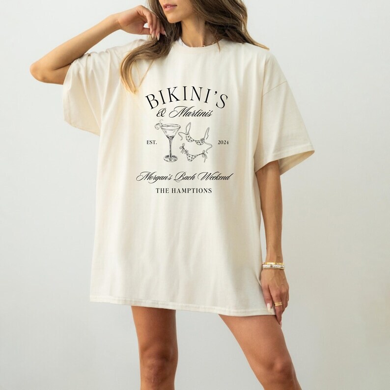 May include: A white t-shirt with a black graphic design that says "Bikini's & Martinis" with a martini glass and bikini illustration. The text also says "Est. 2021" and "Morgan's Bach Weekend The Hamptons".