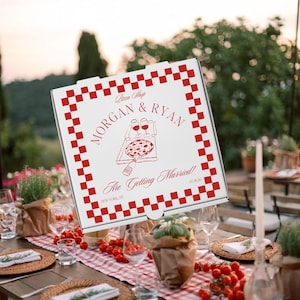 May include: A white square pizza box with a red checkered border. The center features red text reading "Pizza Shop", "MORGAN & RYAN", and "Are Getting Married!", along with a line drawing of a pizza box, two wine glasses, and a bottle, plus "NEW YORK, NY" and "07.30.26".